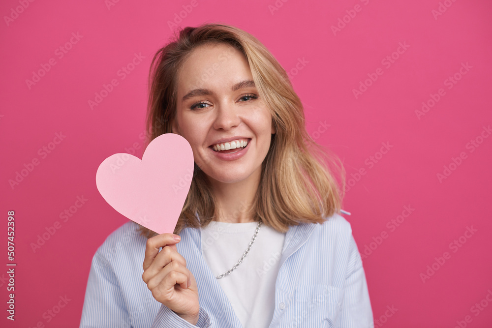 Medium close-up portrait of cheerful Caucasian woman wearing casual outfit standing against pink background holding paper heart, smiling at camera Medium close-up portrait of cheerful Caucasian woman wearing casual outfit standing against pink background holding paper heart, smiling at camera