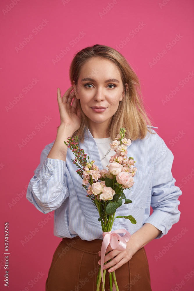 Vertical medium portrait of elegant young woman standing against pink wall background holding beautiful flower bouquet fixing her hair Vertical medium portrait of elegant young woman standing against pink wall background holding beautiful flower bouquet fixing her hair