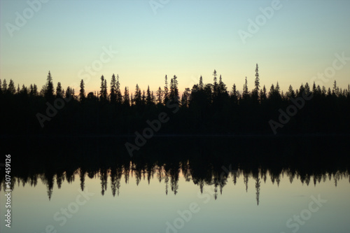 A fantastic view over a northern virgin forest with pines and spruce reflected in the lake during the sunset. Natural colors from gold ocher to light blue in the summer dusk. 