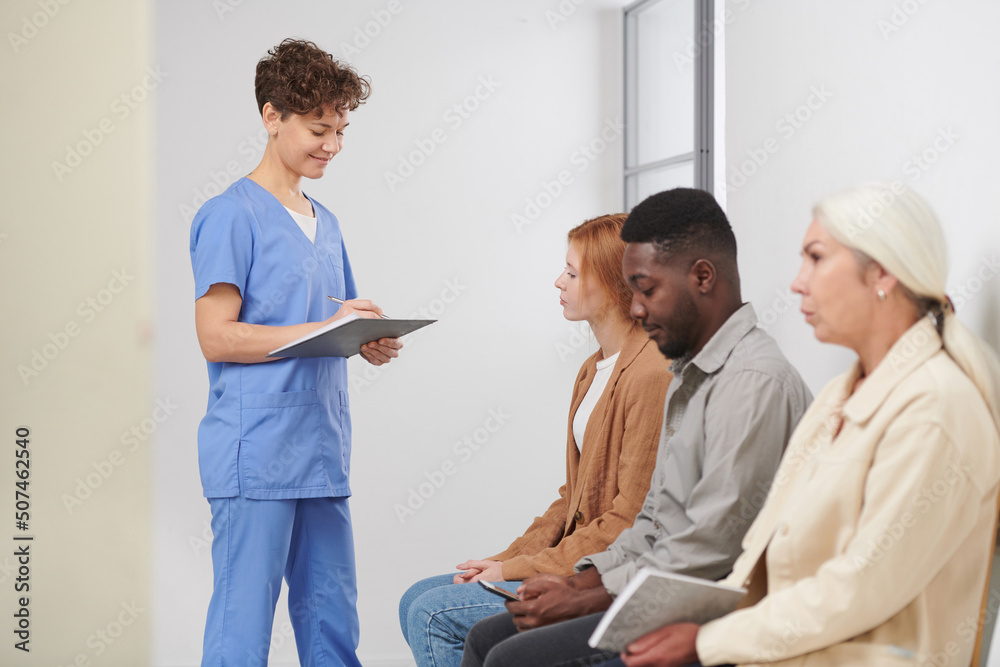 Group of three multi-ethnic people sitting in queue waiting for consultation in modern hospital, medical worker standing near checking attendance Group of three multi-ethnic people sitting in queue waiting for consultation in modern hospital, medical worker standing near checking attendance