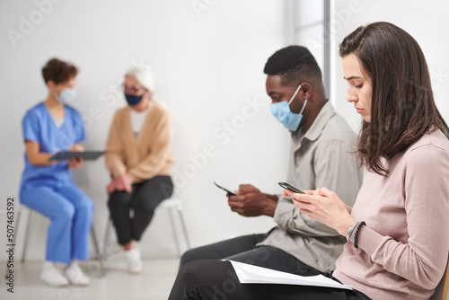 Horizontal shot of man and woman sitting on chair in hospital corridor waiting for consultation time surfing Internet on smartphone, doctor talking to mature woman on background