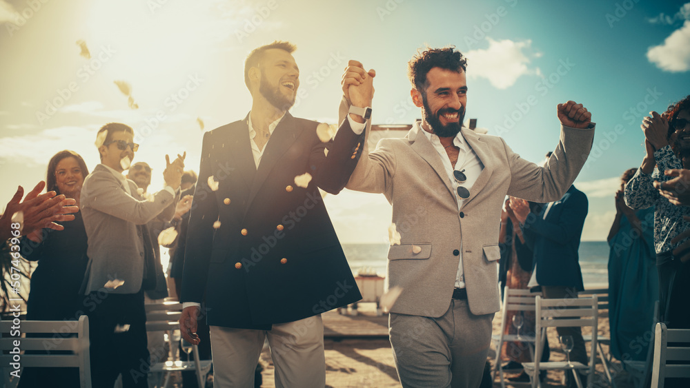 Fototapeta premium Handsome Gay Couple Walking Up the Aisle at Outdoors Wedding Ceremony Venue Near Sea. Two Happy Men in Love Share Their Big Day with Diverse Multiethnic Friends. Authentic LGBTQ Relationship Goals.