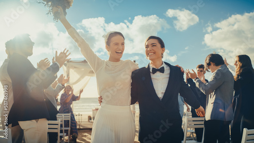 Beautiful Female Queer Couple Walking Up the Aisle at Outdoors Wedding Ceremony Near Sea. Two Lesbian Women in Love Share Happiness with Diverse Multiethnic Friends. Authentic LGBTQ Relationship Goals