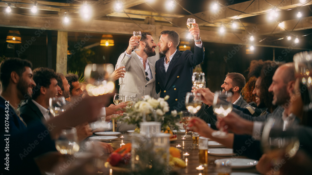 Big Dinner Party with a Crowd of Multiethnic Diverse Friends Celebrating at a Restaurant. Handsome Happy Hosts Propose a Toast and Raise Wine Glasses while Sitting at a Table in the Evening.