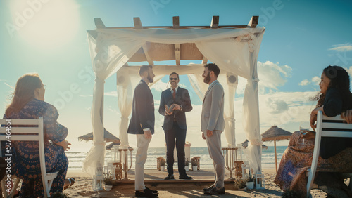 Handsome Gay Couple Exchange Rings and Kiss at Outdoors Wedding Ceremony Venue Near the Sea. Two Happy Men in Love Share Their Big Day with Diverse Multiethnic Friends. LGBTQ Relationship Goals.