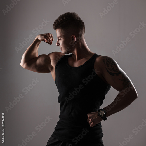 Sporty handsome bodybuilder man model in black fashionable t-shirt shows muscular biceps in dark background