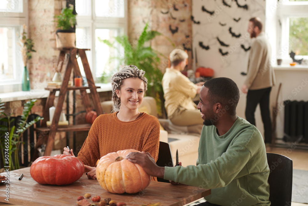 Fototapeta premium Cheerful young interracial friends in sweaters sitting at table in lift studio and carving pumpkins for Halloween party