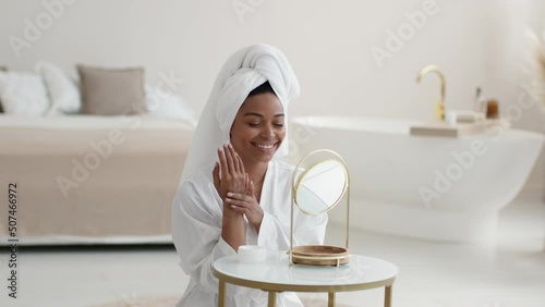 Young pretty african american lady with towel on head applying moisturizing cream on hands, pampering body after shower photo