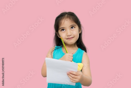 Little Asian girl having paper and pencil in hands looking up, writer waiting for muse, isolated on pink background