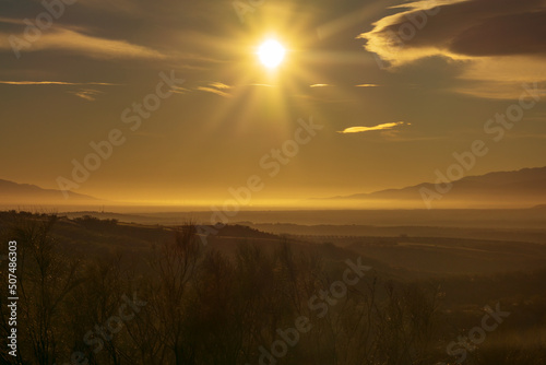 Landscape of fields and low mountains with mists, with a star-shaped sun giving a warm color of dawn to the scene.