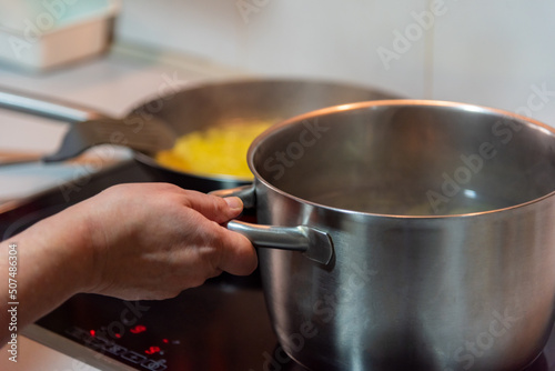 A woman s hand holding a pot that is boiling on a ceramic hob  preparing food.