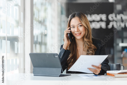 Young brunette Asian businesswoman talking on mobile phone with customer and looking to camera, holding work sheet paper.