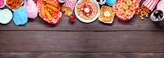 Carnival theme food top border over a dark wood banner background. Overhead view with copy space. Summer fair concept. Corn dogs, funnel cake, cotton candy and snacks.