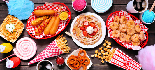 Carnival theme food table scene over a dark wood banner background. Top down view. Summer fair concept. Corn dogs, funnel cake, cotton candy and snacks.