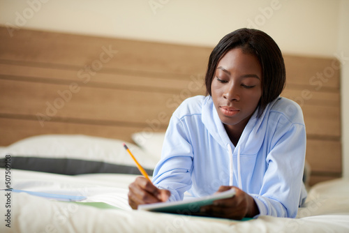 College student lying on bed in dormotory and writing essay in notebook