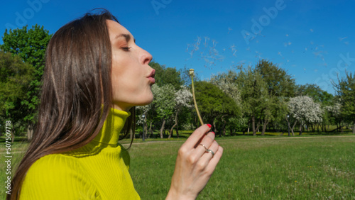 Beautiful young woman blowing a dandelion. Girl with dandelion in hand. Allergic to pollen of flowers. Spring allergy.