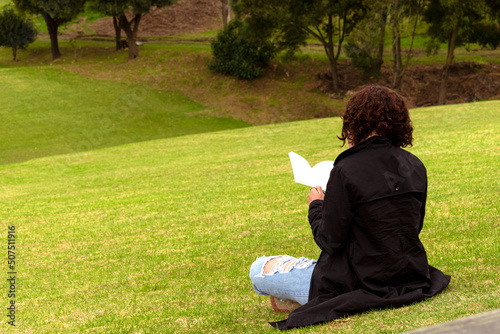 Redhead woman with black sitting on the grass reading a book in a forest