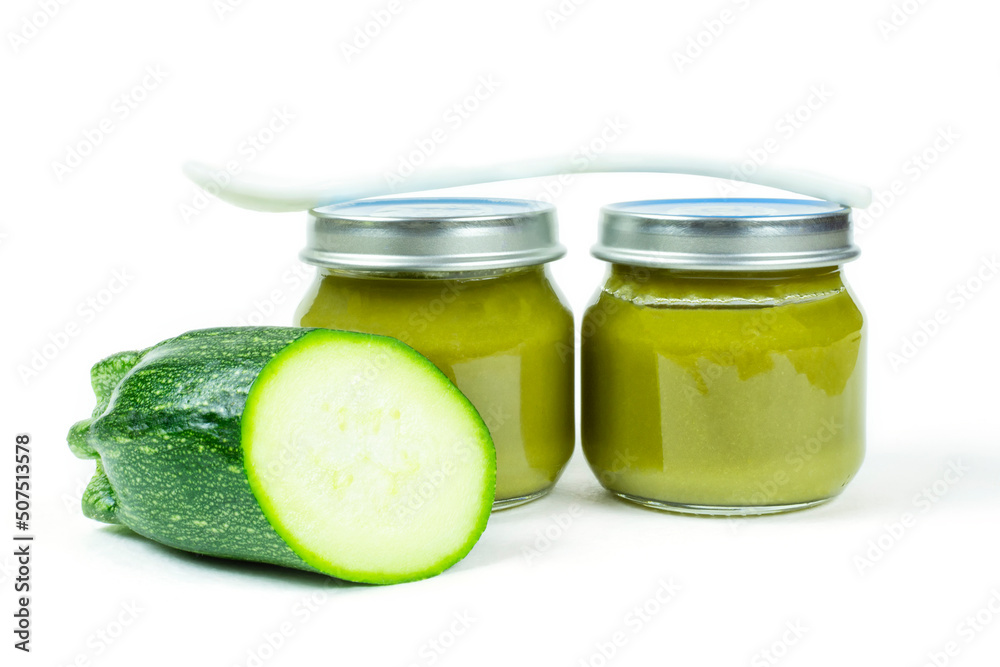 Two glass jars with lids of baby squash puree (zucchini) and a spoon on a white insulated background. Baby food. The first lure of the baby. Two glass jars with lids of baby squash puree (zucchini) and a spoon on a white insulated background. Baby food. The first lure of the baby.