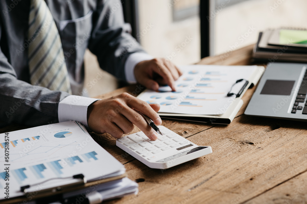 A businessman is using a white calculator to calculate financial numbers, he is sitting in his private office, the businessman examines the financial data from the corporate finance chart. A businessman is using a white calculator to calculate financial numbers, he is sitting in his private office, the businessman examines the financial data from the corporate finance chart.