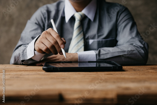Person holding pen pointing at tablet placed on desk, business man looking at information on tablet and using tablet to communicate with company team through messaging and email programs.