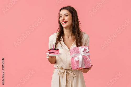 Celebration, party and happiness concept. Carefree smiling happy european woman having fun parying own b-day party, holding gift and piece cake, blow-out candle, pink background