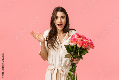 Surprised and happy, excited pretty young woman receive flowers and looking astonished by romantic nice gesture, didnt expact feeling touched raise hand smiling, hold roses, pink background photo