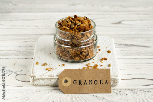 Granola in the glass jar on the white wooden serving board. High key. Healthy ready-ti-eat breakfast. Tag with sign granola