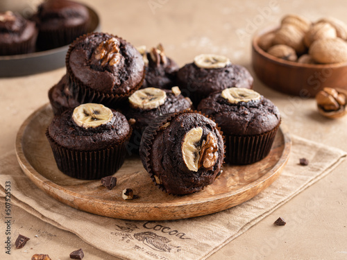 Chocolate banana muffins with chocolate pieces and chopped walnuts on wooden plate. Beige background, textile napkin. Close up food. Homemade brownie dessert. 