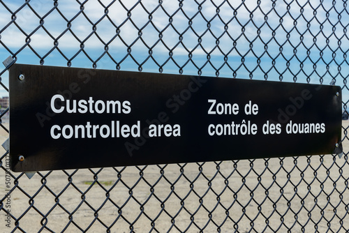 A bilingual Customs Controlled Area sign in English and French on a chain link fence. Angled view with blue sky above, parking lot below.