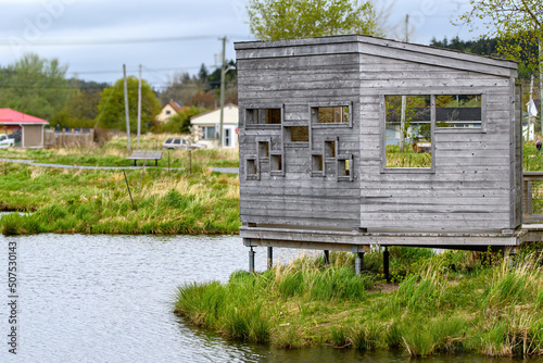 An elevated wooden duck blind beside a pond in a park. Many rectangular holes to look through. Residential area behind, overcast sky.