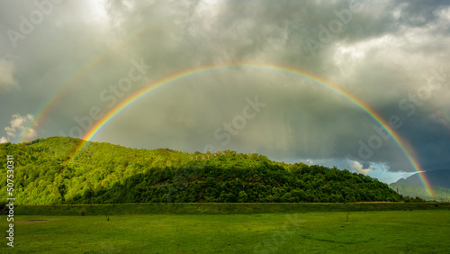 An arched, colorful rainbow appearing above a rural area during a rainy day. The forest is lit by the colorful phenomenon and the sunlight that appears through the stormy clouds. Carpathia, Romania.