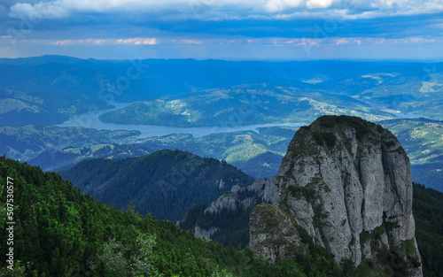 A massive calcareous cliff rising above the wild spruce forests of Ceahlau Mountains. Sun lights the blue waters of a lake and the rural hills below the mountainous landscape. Carpathia  Romania.