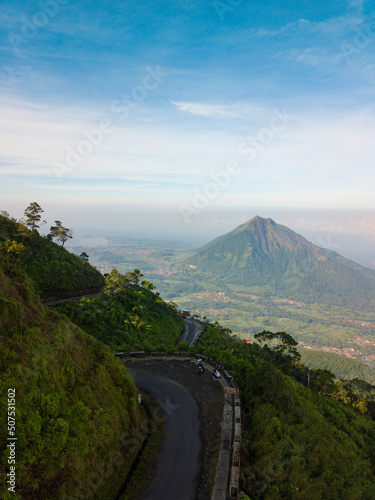 Drone photo of the road on the slopes of a mountain and surrounded by dense forest with Andong Mountain on the background. It located in Mount Telomoyo in clear weather, central java, Indonesia photo