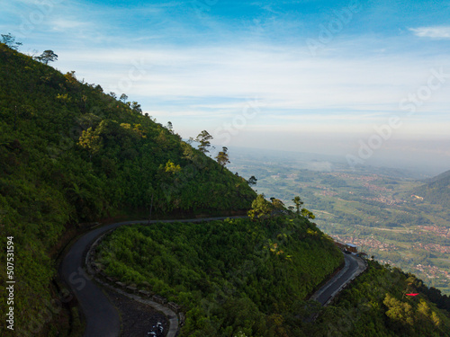 Drone photo of the road on the slopes of a mountain and surrounded by dense forest with Andong Mountain on the background. It located in Mount Telomoyo in clear weather, central java, Indonesia