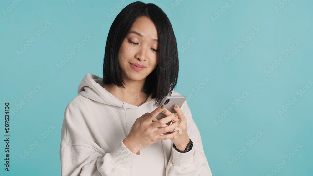 Attractive Asian woman wearing white hoodie chatting with boyfriends using smartphone on blue background Attractive Asian woman wearing white hoodie chatting with boyfriends using smartphone on blue background