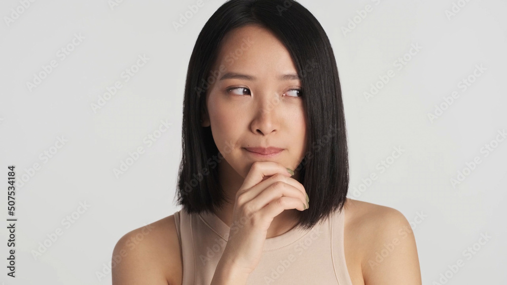 Asian woman slyly looking around keeping hand on chin and thinking over white background. Female thinking out a plan Asian woman slyly looking around keeping hand on chin and thinking over white background. Female thinking out a plan