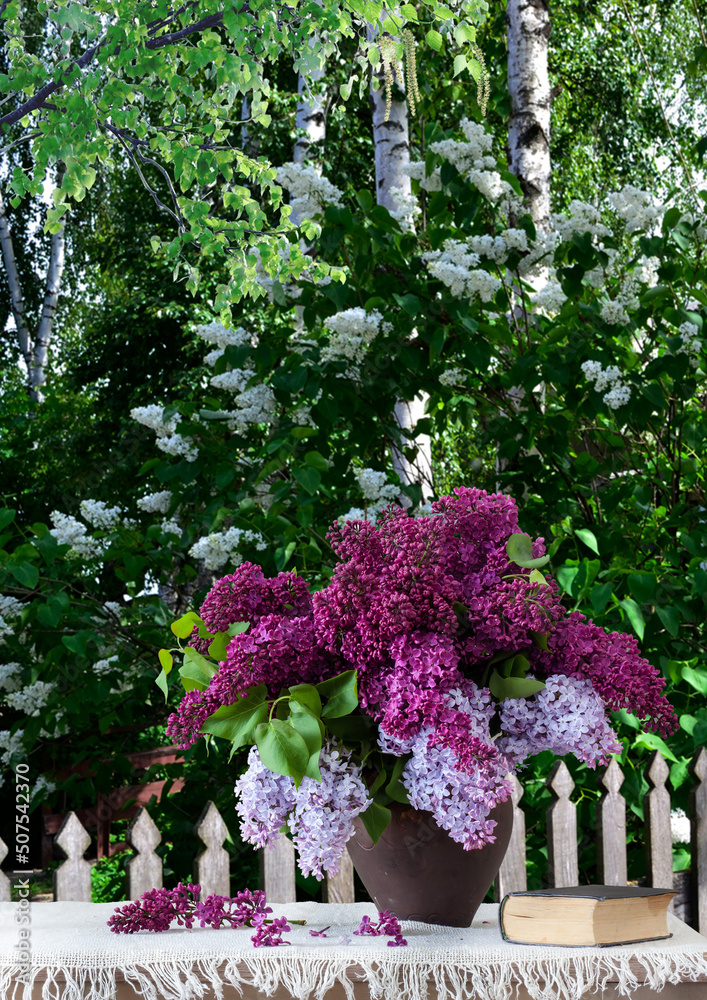 bouquet of lilacs in a clay jug on a wooden table with a white tablecloth against the background of slender birches background for decoration and design