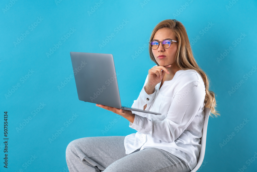 Beautiful attractive young business woman holding laptop computer in hands, posing angry with pain in her face, in white shirt and grey pants. Isolated over blue background.