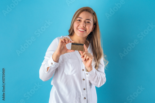 Beautiful attractive young business woman posing happily while holding credit card, in white shirt and grey pants. Isolated over blue background.
