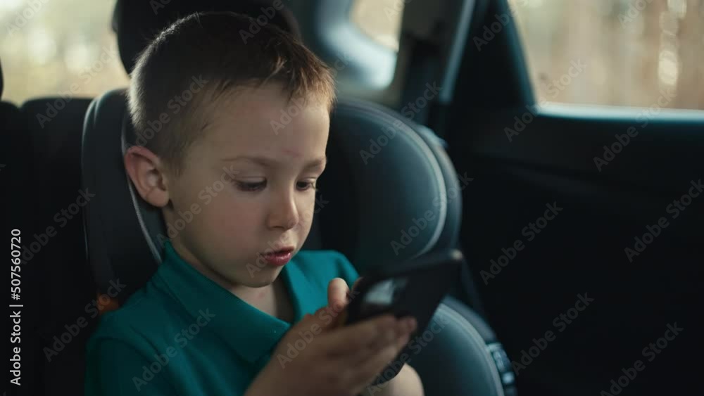 A child plays games on the phone while traveling by car. A boy sitting in a child car seat with a gadget while traveling with his parents. A child plays games on the phone while traveling by car. A boy sitting in a child car seat with a gadget while traveling with his parents.