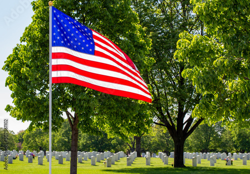 Close up view of an American flag in a military cemetery setting with trees and blue sky and with a light breeze