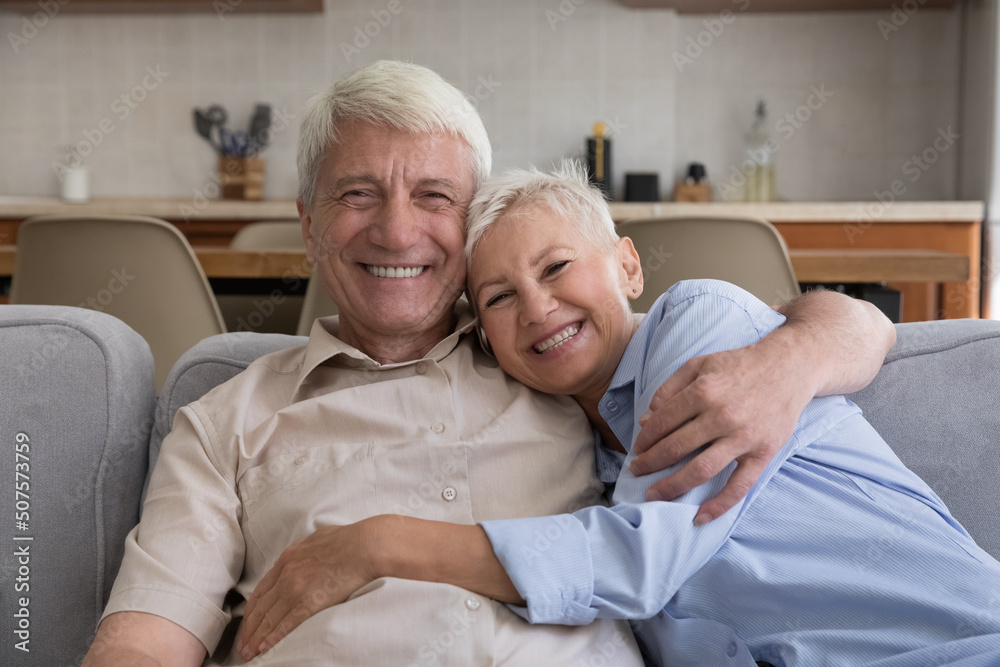 Headshot family portrait of two happy pensioners hoary senior adult husband wife cuddling on sofa. Loving family couple of grandparents laughing hugging looking at camera with healthy bright smiles Headshot family portrait of two happy pensioners hoary senior adult husband wife cuddling on sofa. Loving family couple of grandparents laughing hugging looking at camera with healthy bright smiles
