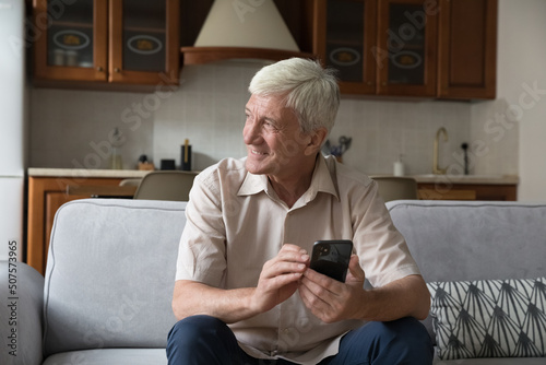 Happy casual senior adult male staying at home use phone for online communication send receive message in chat look at distance think about good news. Smiling aged man spend free time with modern cell