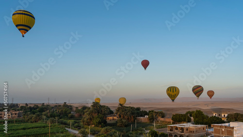 Bright balloons are flying in the blue sky. Below you can see rural houses, palm trees, cultivated fields. In the distance - the sand dunes of the desert. Egypt. Luxor