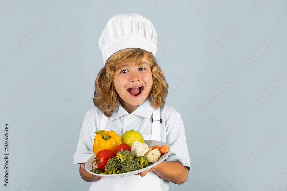 Excited funny chef cook. Portrait of a 7, 8 years old child in cook cap and apron making fruit salad and cooking food in kitchen. Cute little blonde happy smiling chef hold plate with vegetables. Excited funny chef cook. Portrait of a 7, 8 years old child in cook cap and apron making fruit salad and cooking food in kitchen. Cute little blonde happy smiling chef hold plate with vegetables.