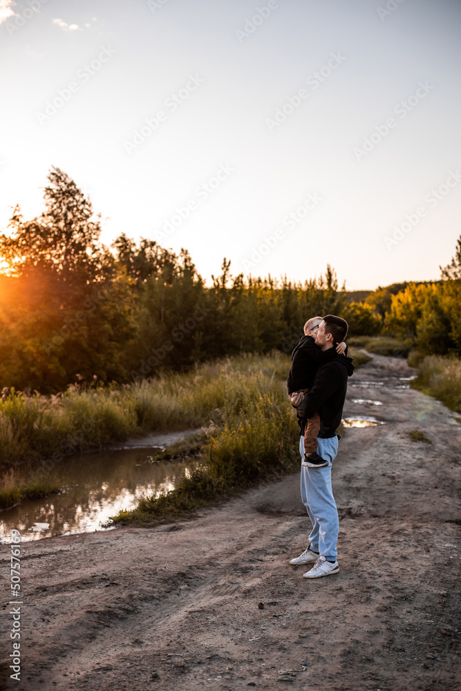 Fototapeta premium Dad and son walk in nature in the park at sunset along a country road. The father holds his son in his arms. The son is happy and happy. Happy family moments.