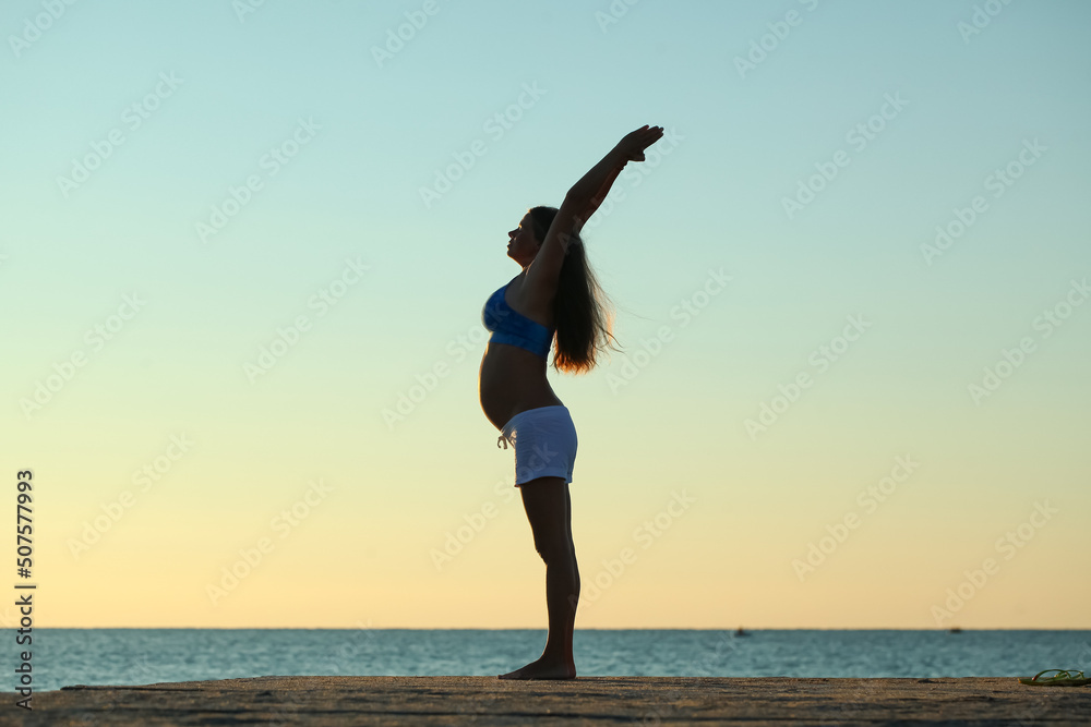 Obraz premium pregnant girl doing yoga on the seashore in the morning against the sky and water