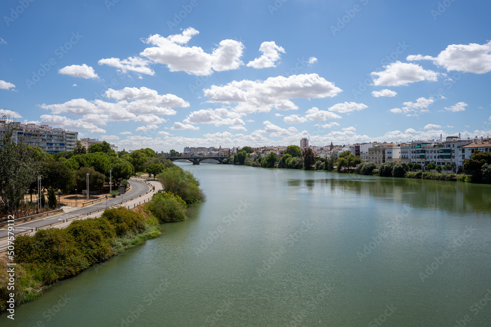 a large wide green river that flows through the Spanish city of Seville with green trees and white houses on both sides