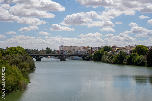 a large, wide green river that flows through the Spanish city of Seville with green trees and white houses on both sides, and an old stone bridge in the distance