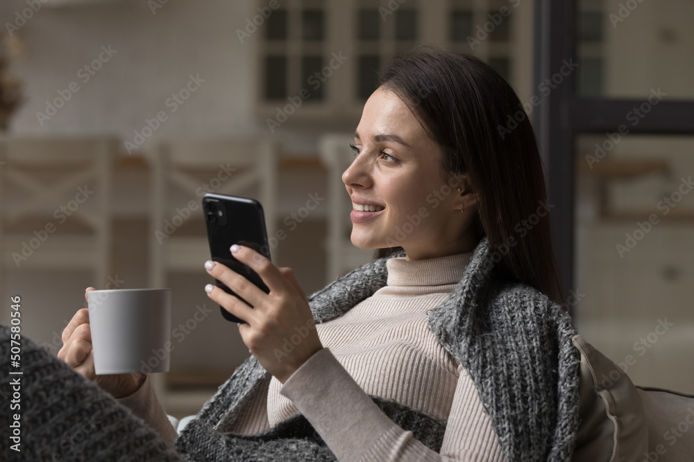 Thoughtful happy mobile phone user girl wrapped in warm cozy plaid holding mug of hot drink, morning coffee, tea, enjoying leisure at home in winter, autumn time, thinking, dreaming